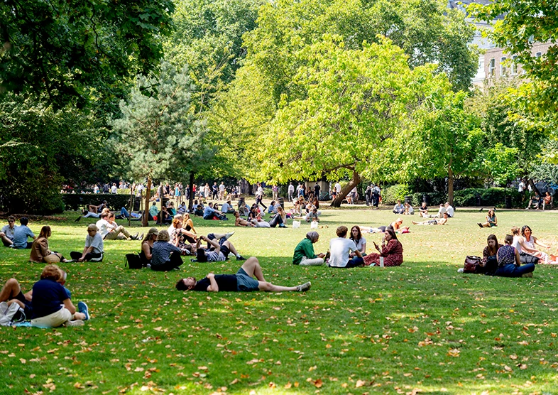 Lincoln’s Inn Fields is a lively park pictured on a
sunny day with people relaxing on the grass,
sitting in groups, or lying down under the shade
of large green trees. The park is filled with lush
foliage and dappled sunlight, creating a vibrant
and peaceful atmosphere. 