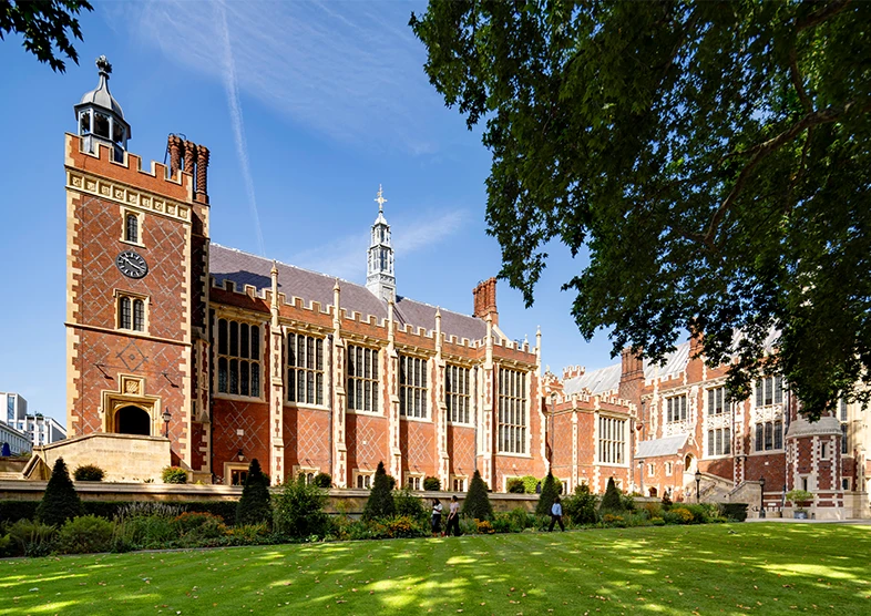 Historic red-brick building which is Lincoln’s Inn
in London, featuring Gothic architecture, large
windows, and landscaped gardens under a clear
blue sky.
