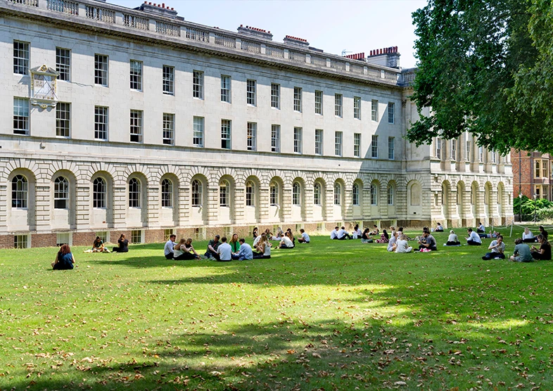 Stone building and walkways can be found on
Lincoln’s Inn Estate. People sit to eat lunch on
the well-maintained green lawn in front of the
buildings, there are multiple leafy trees that
provide shade.