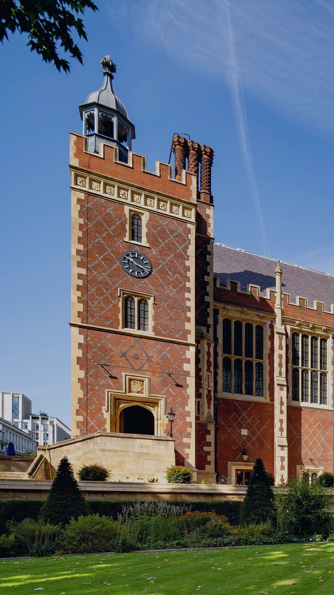 Historic red-brick building which is Lincoln’s Inn
in London, featuring Gothic architecture, large
windows, and landscaped gardens under a clear
blue sky.
