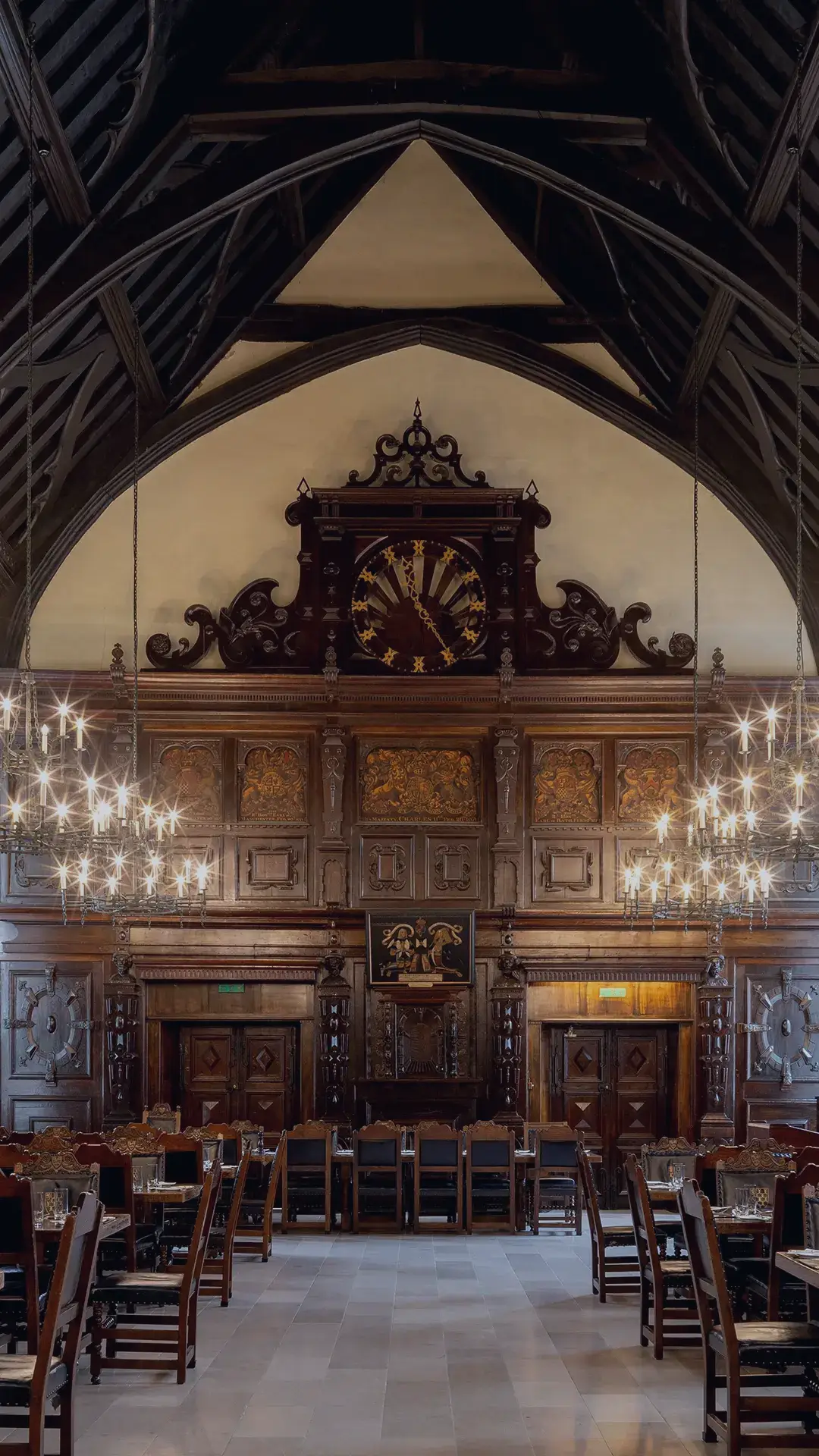 The Old Hall is a grand, historic dining hall with
high vaulted ceilings and dark wooden beams.
The room is lined with long wooden tables set
with silverware and glassware. Ornate
chandeliers hang from the ceiling, casting warm
light. Stained glass windows and large oil
paintings decorate the stone walls, and an
intricately carved wooden gallery and organ are
visible at the far end of the hall.
