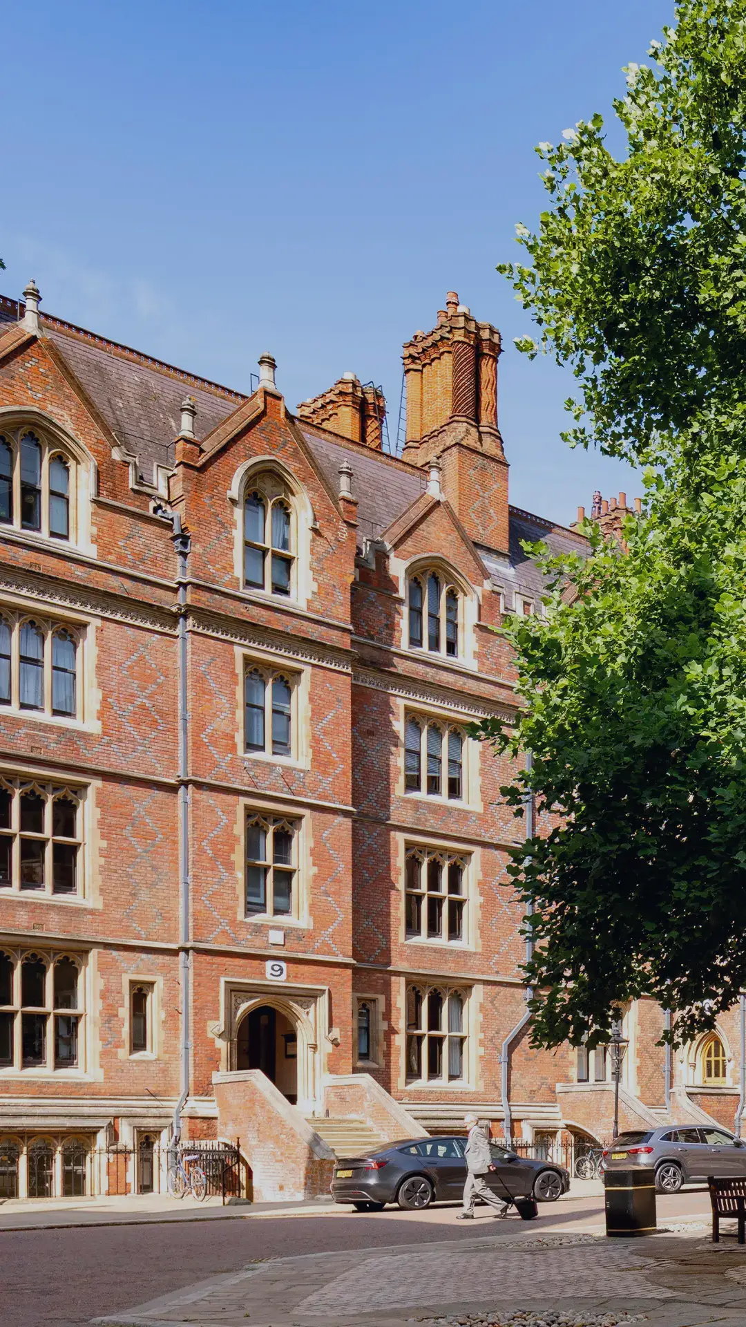Old Square is a central courtyard located on
Lincoln’s Inn Estate, a large leafy tree is in the
centre and is surrounded by red-brick Gothicstyle buildings and parked cars. Benches circle
the tree under a clear blue sky.
